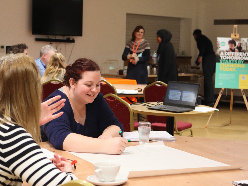 Photograph of two people sat at a table, both smiling on writing on a large sheet of paper.