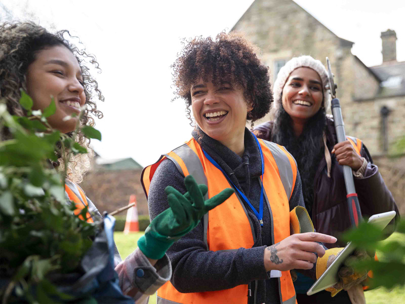 Three women in high-visibility vests, smiling and talking while holding gardening tools in an outdoor setting.