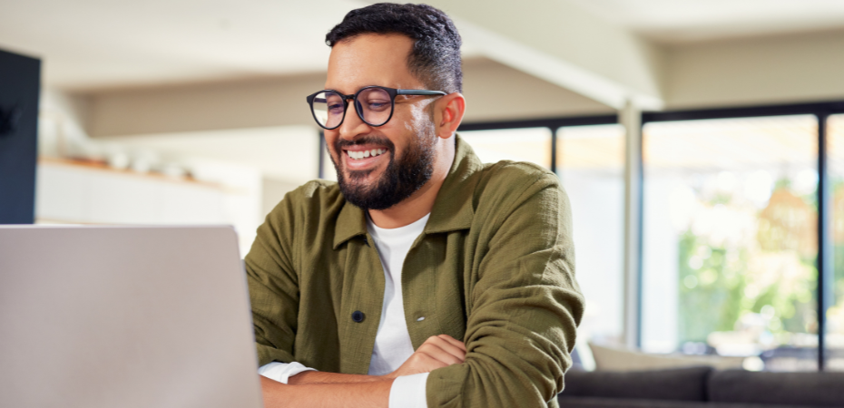 Photogrpaph of a man sitting at a table using a laptop, he wearing black framed glasses and a green shirt. His arms are folded and he is smiling.