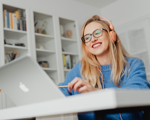 Photograph of a young woman using a laptop, she is smiling and wearing bright orange earphones.