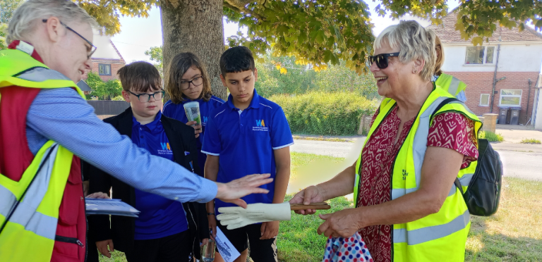 A photograph of a man, wearing a high visibility vest handing an artefact to a woman in a high visibility vest. They are in a part and overlooked by three young school boys.