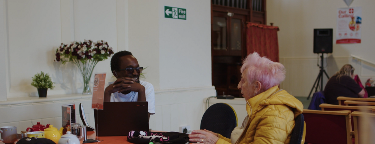 A photo of a Digital Champion sat at a table with a laptop in front of him, engaging in conversation with a woman wearing a yellow coat.