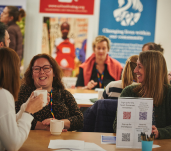 A group of women sat at a table at an event. They have mugs of tea and coffee with them, and are smiling and laughing with each other.