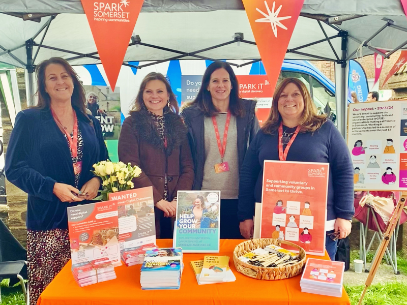 A photo of four members of the Spark Somerset team, posing at a stand featuring leaflets and posters from our organisation's projects. They are smiling at the camera.