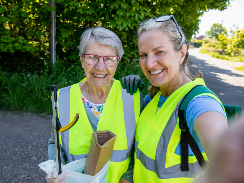 A selfie-style photo of two women wearing fluorescent vests and holding litter-pickers, standing on the side of a country road. They are smiling at the camera.