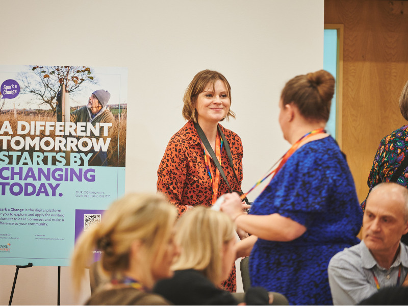 A photo of a member of the Spark Somerset team next to a board advertising Spark a Change. She is smiling at a woman who has her back turned to the camera.