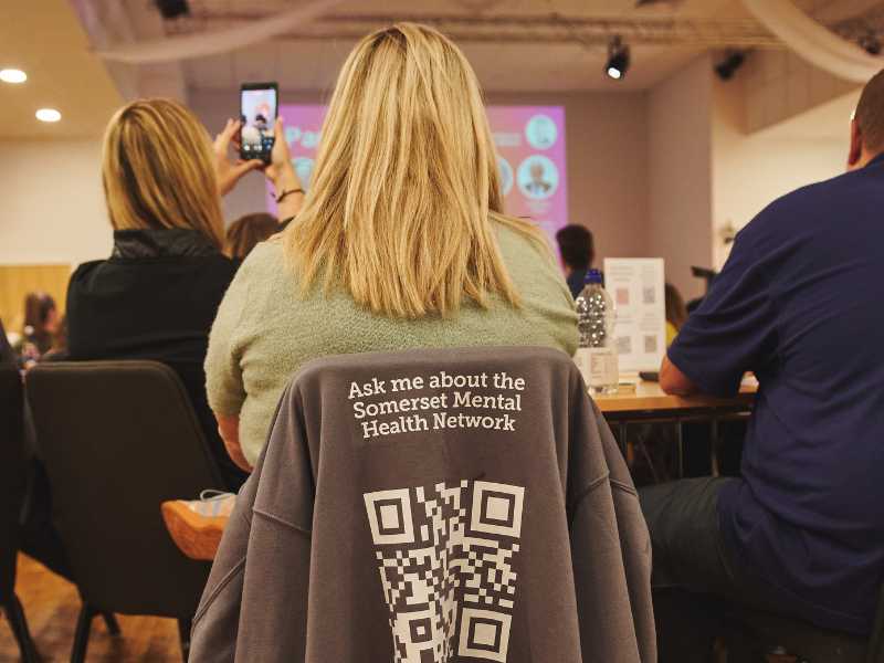 A photo of a member of the Spark Somerset team. She is sat with her back to the camera. On the back of her chair is a fleece that says 'Ask me about the Somerset Mental Health Network' and features a QR code