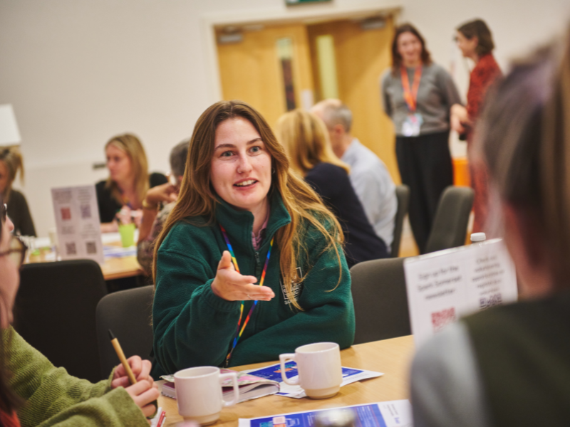 A photo of a woman in a green fleece, sat at a table and engaging in conversation with the people sat around her