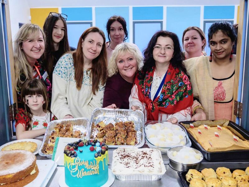 A photo of a group of women, each wearing clothing from their cultural background. There are trays of various baked goods in front of them.