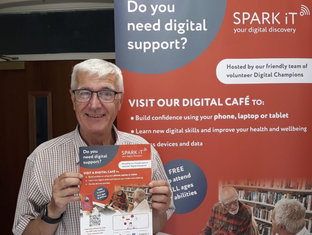 A photo of a man posing next to a Spark iT pull-up banner, and holding a Spark iT flyer. He is looking at the camera and smiling.