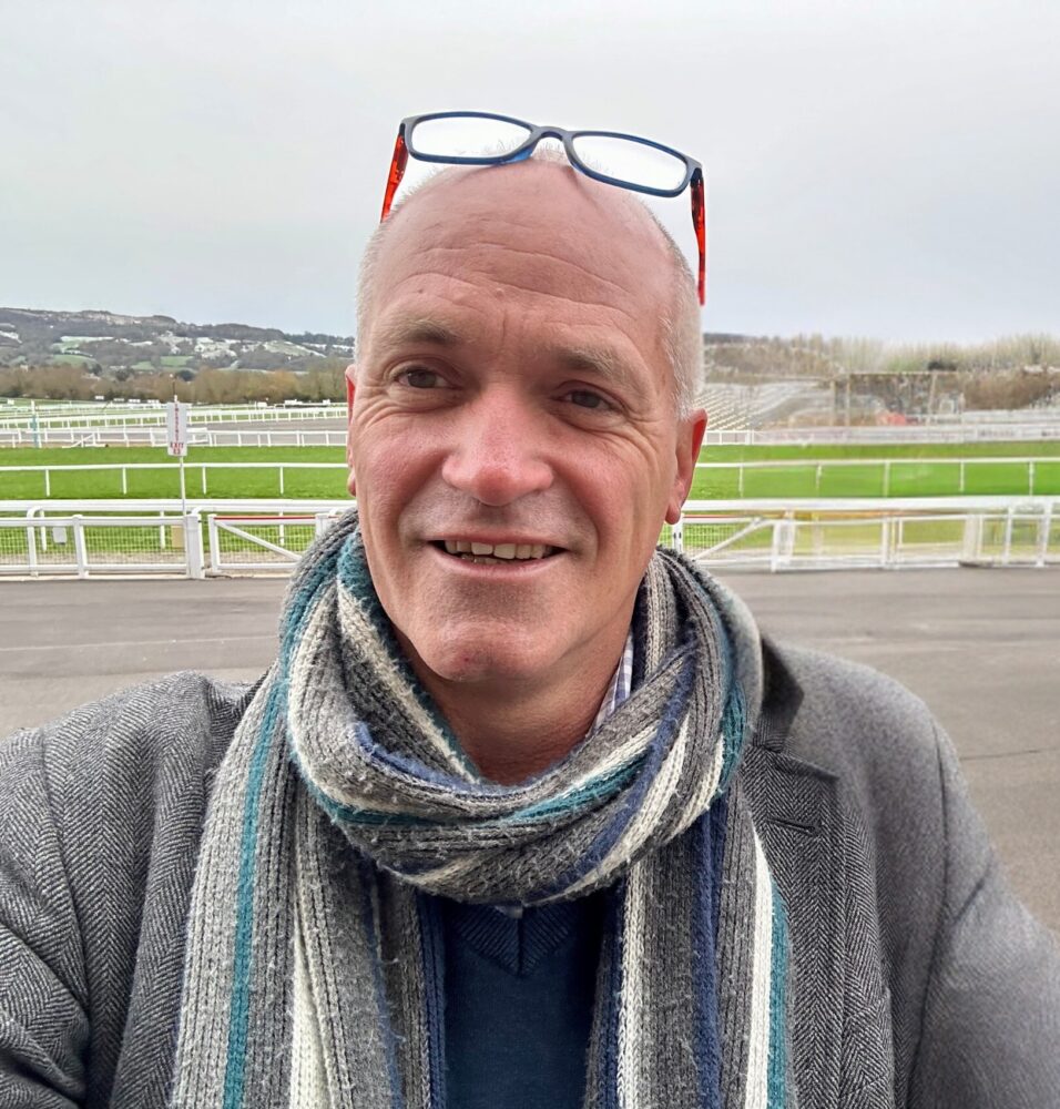 A photo of Nik Harwood. He is wearing a scarf and a grey wool jacket, and smiling at the camera