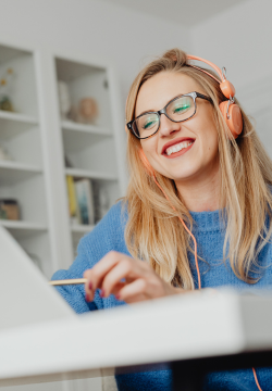A photo of a woman sat at a desk. She is wearing headphones and glasses and is smiling down at the laptop in front of her.