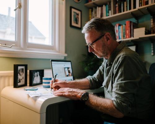 Photo of a man with grey hair and glasses, sat at a desk and filling in a form