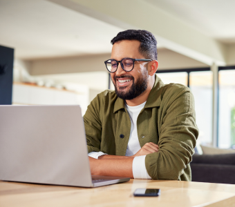 Picture of a man sat at a table in front of a laptop. He is wearing glasses and a green shirt, and is smiling at the laptop.