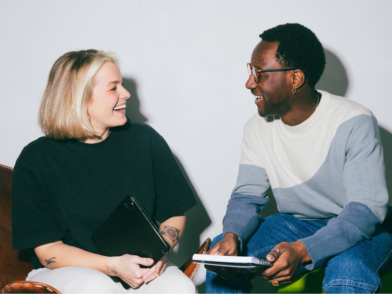 Photograph of a man and a woman sitting next to and smiling at each other. The woman is holding a laptop and the man is holding a notepad. Plain white wall in the background.
