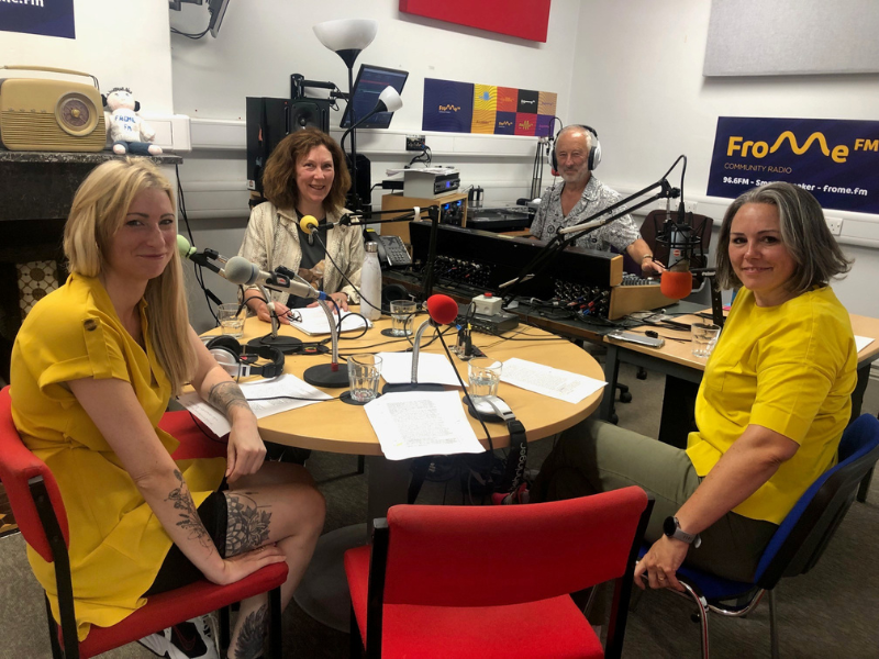 A photo of a member of the Spark Somerset team sat alongside members of the Frome FM Community Radio team. They are in the radio studio, with microphones in front of them.