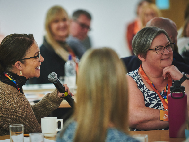 Photograph of three women and an event, one is speaking into a microphone, one has her back to the camera and one is looking away and smiling.