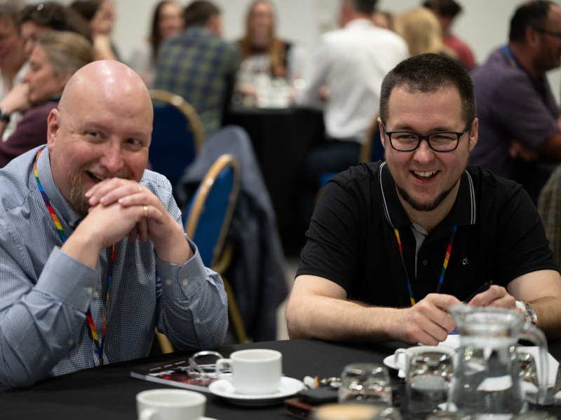 A photo of two men sat at a table, with glasses of water and coffee cups in front of them. They are looking to someone beside the camera, and laughing.