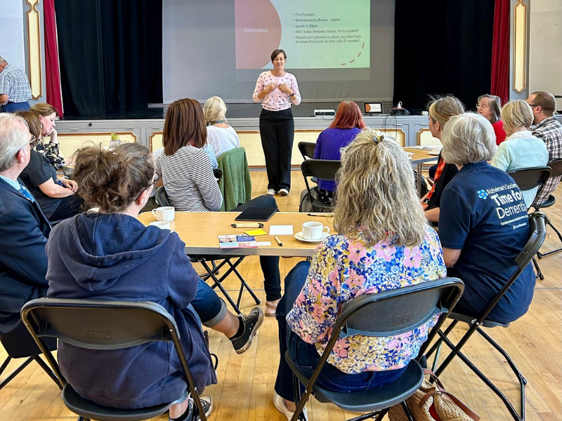 Photograph of people sitting at tables in a community hall, they have their backs to the camera and are looking at a woman presenting in front of a large screen.