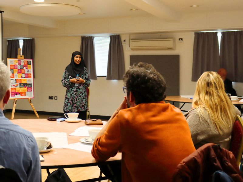 A photo of a lady wearing a hijab, presenting to a room of people sat at tables.
