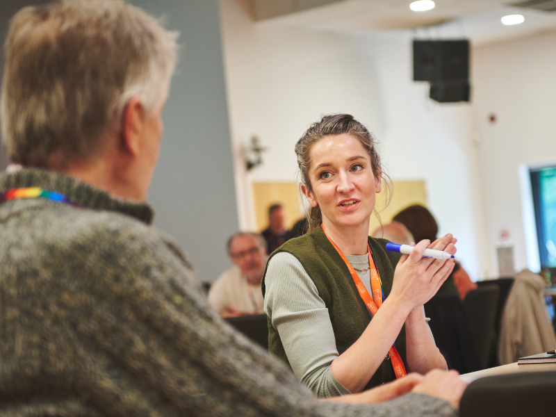 A photo of a woman in mid-conversation with a man sat in front of her. They are both sat at a table and she is holding a pen.