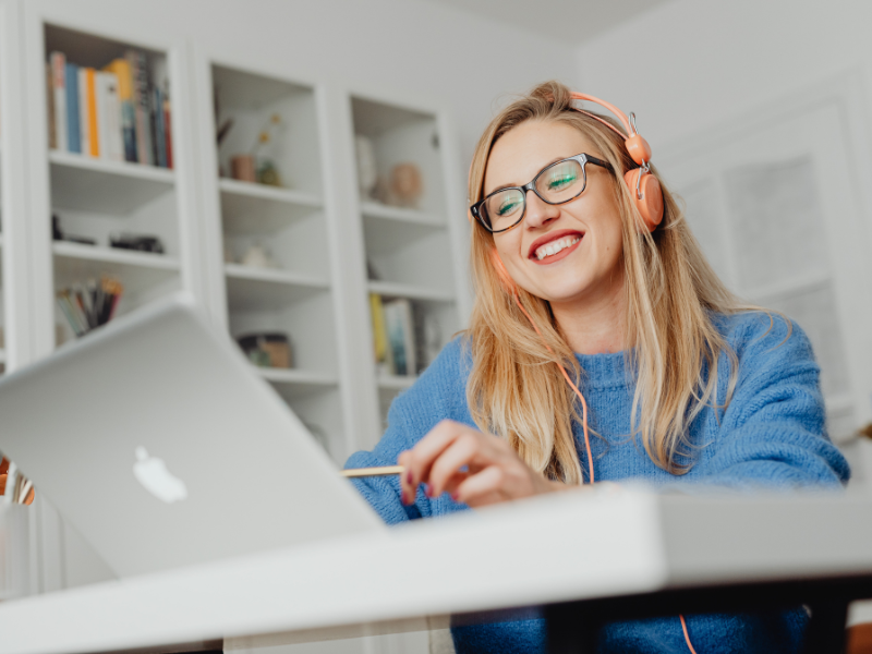 Photograph of a young woman smiling and using a laptop, she is wearing a blue jumper and orange headphones.