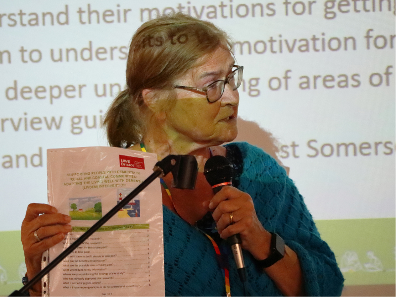 Photograph of a woman, close up speaking into a microphone and standing in front of presentation being projected onto, and behind her.