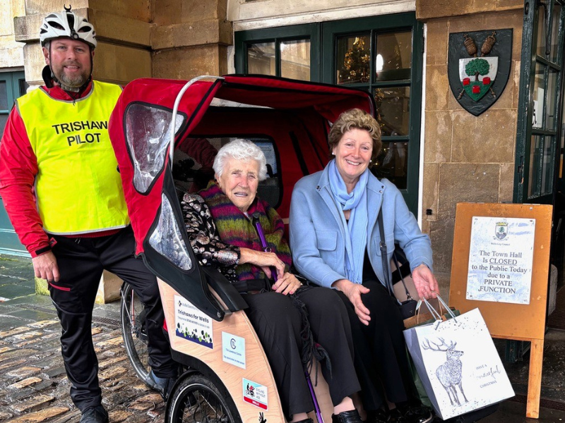 Photograph of two women sat in a trishaw, the are smiling at the camera and one is holding a shopping bag. The trishaw cyclist is behind them, also smiling and wearing a hi-vis vest with 'Trishaw Pilot' written on it.