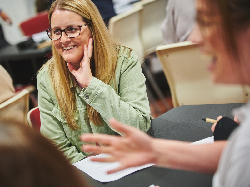 Photo of a woman with her elbow propped on a table, listening to a conversation and smiling