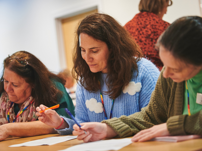 A photograph of three women sat at a table writing on some paper, the woman in the middle has a pale blue jumper with a cloud pattern.