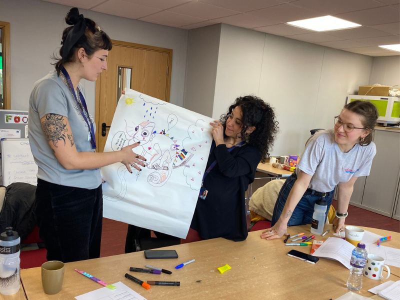 A picture of three young people stood near a table. Two of them are holding a piece of paper with a drawing of a chameleon on it. The table in front of them is covered in marker pens.