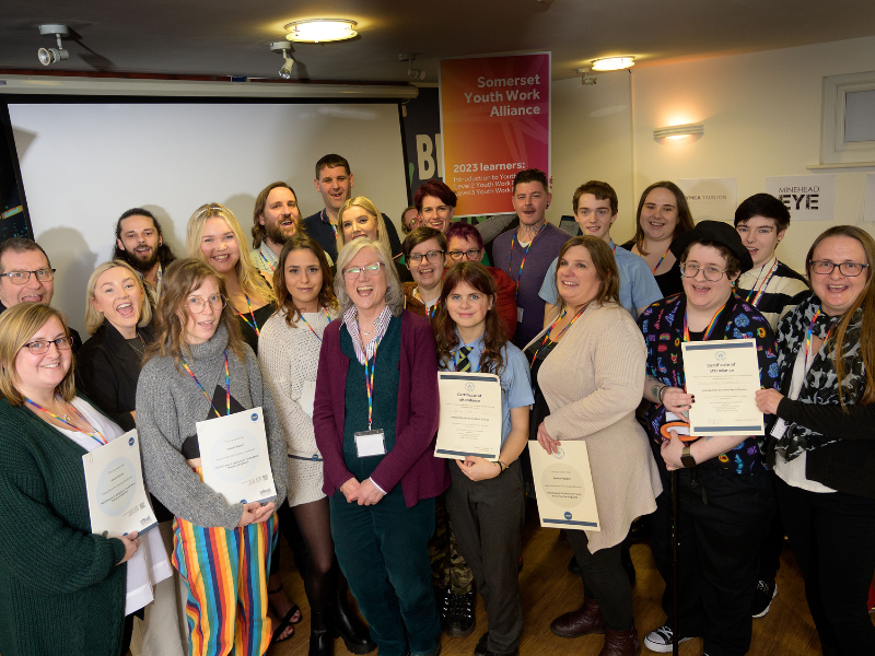 A large group of young people are stood together, holding certificates of achievement, and looking up and smiling at the camera.