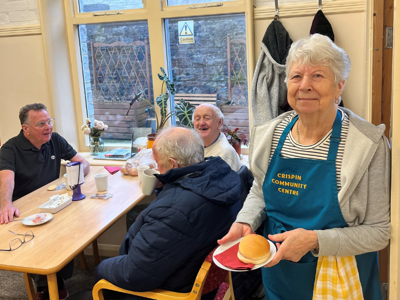 Photograph of three men sitting at with hot drinks. One is smiling at the camera. In the foreground a woman is smiling at the camera holding a bacon roll and wearing an apron with the words 'Crispin Community Centre'.