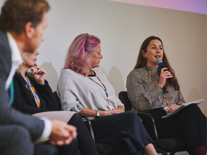Photograph of four people sat in a row on stage at an event. One woman is speaking into a microphone, the others are looking at her.