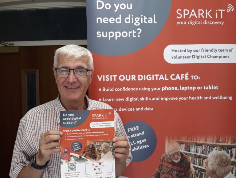 A photo of a man posing next to a Spark iT pull-up banner, and holding a Spark iT flyer. He is looking at the camera and smiling.