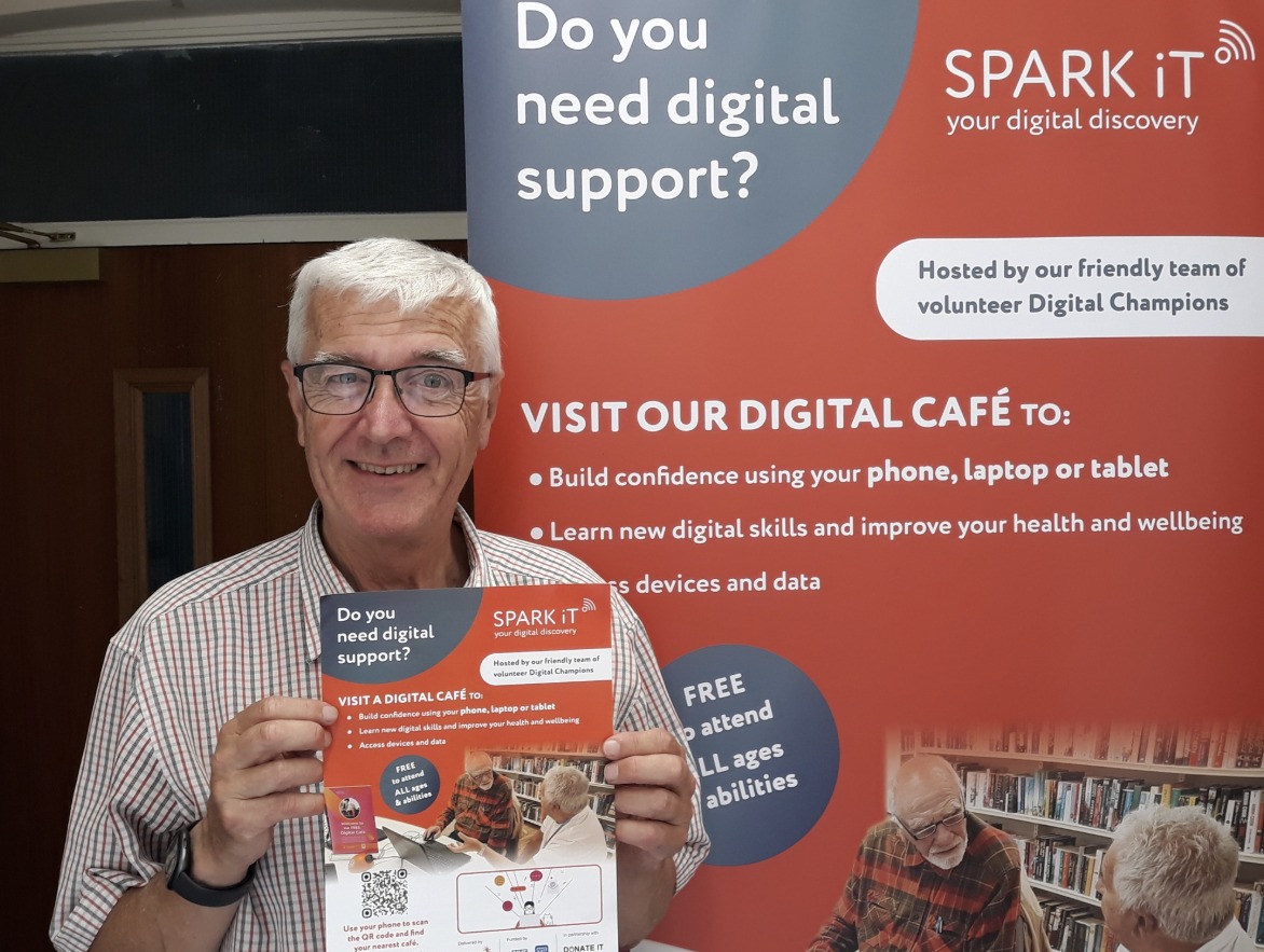 A photo of a man posing next to a Spark iT pull-up banner, and holding a Spark iT flyer. He is looking at the camera and smiling.