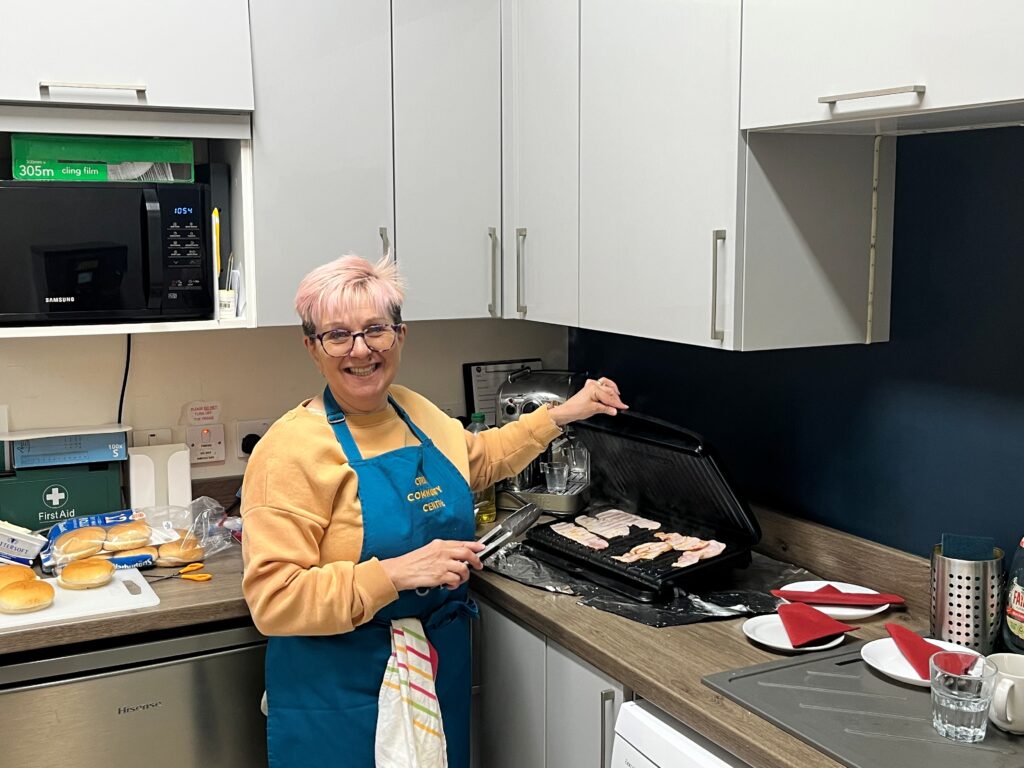 A picture of a Warm Welcome volunteer in a kitchen. She is cooking bacon on a grill, and is looking at the camera and smiling.