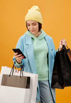 Photograph of a fashionable woman carrying shopping bags and looking at her mobile phone on a bright yellow background.