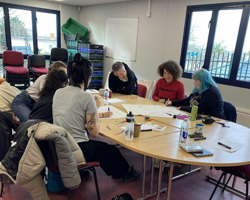 A group of young people in a classroom setting, are sitting around a table with pieces of paper in front of them. They are engaging with each other.