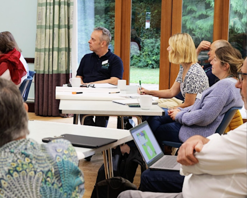 A photo from a Volunteer Coordinators' Forum; people are sat at tables with notepads and laptops and are listening to the host.