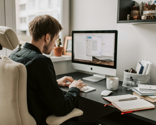 Photo of a young man sat at a computer, with open notebooks near him on the desk. He is photographed from the back, and we can see his screen.