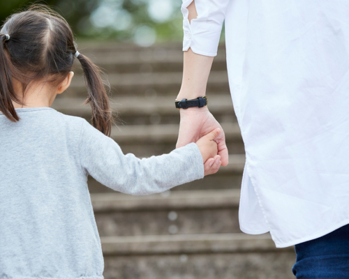 Picture of a child with bunches holding the hand of an adult. They are pictured from behind as they walk up a set of stone steps.