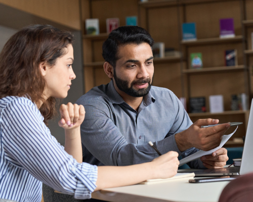 A photo of a man and a woman sat at a laptop. They have pieces of paper in front of them, and are in discussion.