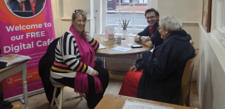 Lynne, featured in the article sitting at a table with a young male volunteer and older female client. They are smiling at the camera. There is a Spark iT promotional banner behind Lynne.