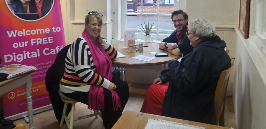 Lynne, featured in the article sitting at a table with a young male volunteer and older female client. They are smiling at the camera. There is a Spark iT promotional banner behind Lynne.