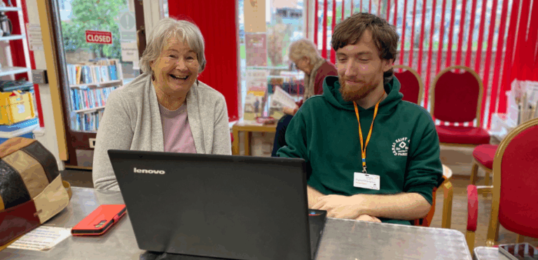 Sue a visitor to the Digital Cafe featured in the story sitting alongside volunteer Digital Champion Morgan. They have a laptop in front of them on the table and Sue is smiling warmly at the camera.