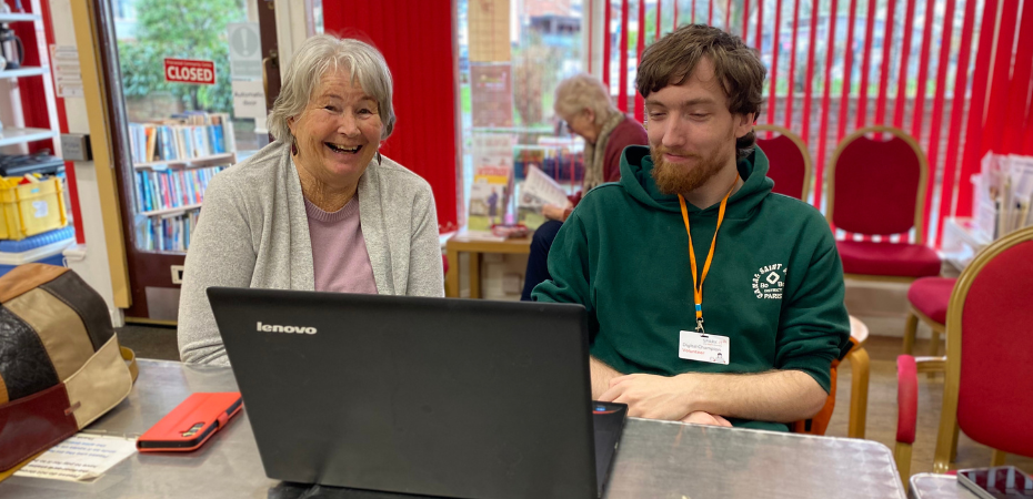 Sue a visitor to the Digital Cafe featured in the story sitting alongside volunteer Digital Champion Morgan. They have a laptop in front of them on the table and Sue is smiling warmly at the camera.