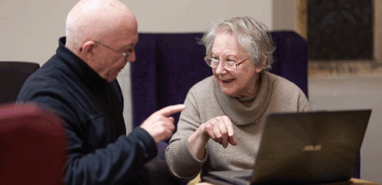 A photo of a man and and older lady sat at a desk with a laptop, engaged in conversations