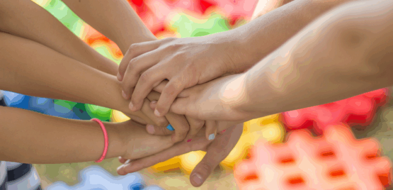 Childrens holding hands in a circle with only their hands and forearms in shot, some have painted fingernails and there are colourful lego type blocks in the background.