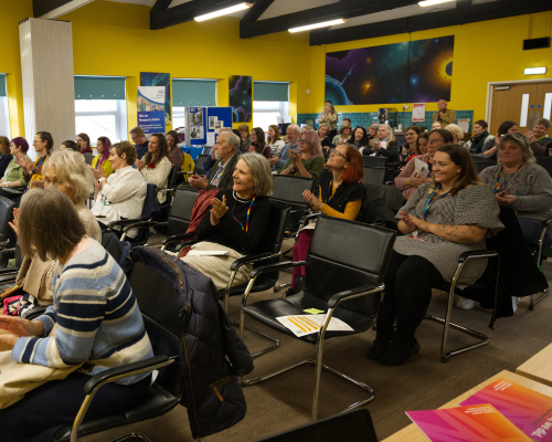 An image from a previous Spark Somerset event; a room full of people are sat listening to a speaker. They are smiling and clapping.